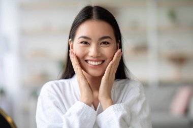 Beyaz bornozlu Asyalı bir kadının cildine dokunup gülümsemesi, pürüzsüz teninin tadını çıkarması, fotokopi çekmesi. Neşeli genç Çinli bayan iki eliyle yanaklarına dokunuyor.