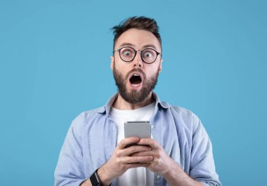 Şok olmuş sakallı adam mavi stüdyonun arka planında cep telefonuna bakıyor. Akıllı telefondan inanılmaz haberler öğrenen şaşırtıcı genç adam internette büyük bir satış yaptı.
