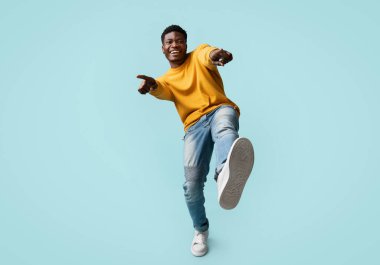 Kaygısız, yakışıklı, komik, bin yıllık siyah adam mavi stüdyo arka planında poz veriyor, kameraya gülüyor ve el kol hareketi yapıyor, Afro-Amerikan genç adam kameraya işaret parmağı ve ayakkabı gösteriyor.