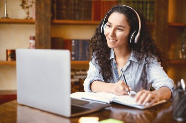 Modern Teknoloji ve Eğitim. Kablosuz kulaklıkla gülümseyen kadın Webinar izliyor, masada oturuyor, dizüstü bilgisayar kullanıyor ve not defteri yazıyor, not alıyor. Karantina Sırasındaki Eğitim