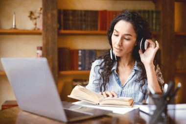 Bilgi kavramı. Sakin, genç bir bayanın portresi kablosuz kulaklık takıp dokunması, kağıt kitap okuması, dizüstü bilgisayarla masaya oturması, evde online çalışması, ödev yapması, ara vermesi.