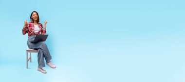 Asyalı kadın koltukta oturuyor, gülümsüyor ve ellerini kaldırıyor. Parlak mavi arka plan onun neşeli anına neşe katıyor. Bir başarı duygusunu yakalıyor..