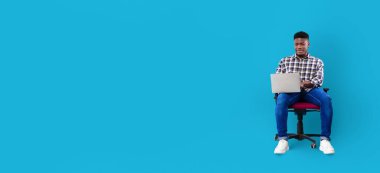 Afro-Amerikalı genç adam bir ofis sandalyesinde rahatça otururken dizüstü bilgisayarına odaklanıyor. Parlak mavi arka plan modern bir dokunuş ekleyerek, işine yoğunlaşmasını vurguluyor..