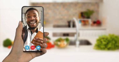 Samimi bir mutfakta, bir adam akıllı telefon ekranında parlak bir şekilde gülümser, bir video görüşmesiyle meşgul olur. Arka plandaki taze sebzeler neşeli bir ev ortamını işaret ediyor..