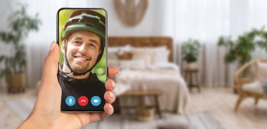 Bir video görüşmesi sırasında gülümseyen bir kişi akıllı telefon ekranında görünür. Arka planda ışık perdeleri ve rahat mobilyaları olan iyi dekore edilmiş bir ev var..
