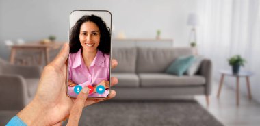 Bir kişi akıllı telefon kullanarak bir video görüşmesine katılıyor, rahat bir ev ortamından gülümseyen bir kadınla iletişim kuruyor. Dekor, rahat mobilyaları olan modern bir oturma odasını içeriyor..