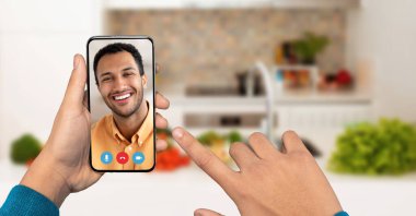 Parlak bir mutfaktaki akıllı telefon kullanarak ekranda başka bir kişinin gülümseyen yüzünü gösteren bir video çağrısına katılan bir kişi. Arka planda taze sebzeler var..