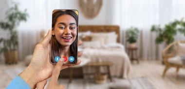 Bir kişi rahat bir odada video görüşmesi yaparken elinde akıllı telefon tutuyor. Arka planda rahat bir yatak ve bitkiler var. Sohbet için sıcak bir ortam yaratıyor..