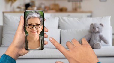 Bir kişi akıllı telefonu evinden gelen bir görüntülü arama için kullanıyor. Ekrandaki kadın gülümsüyor ve yakınlarda bir oyuncak ayıyla kanepede dinlenirken sohbete daldı..