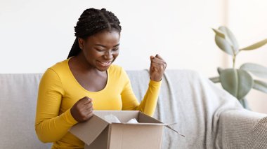 Mutlu siyahi kadın karton kutuları açıyor, başarılı alışverişe duygusal tepki veriyor, büyük alışverişlerle tatmin oluyor, heyecandan yumruklarını sıkıyor, oturma odasında kanepede oturuyor.