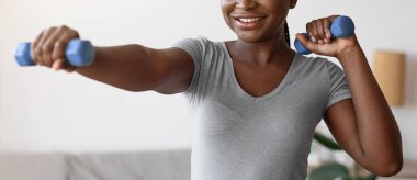 Spor portresi, evde dambıllarla egzersiz yapan siyah kadın, sağlıklı yaşam tarzının tadını çıkaran, ev içi eğitimin keyfini çıkaran, motive olan, uzaklara bakan ve gülümseyen,