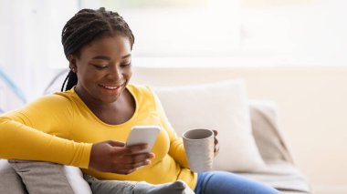 Hafta sonu tatili. Akıllı telefon kullanan ve evde çay içen neşeli siyah kadın, oturma odasında cep telefonu ve kahveyle dinlenen bayan, internette gezinen ya da sosyal ağları kontrol eden,