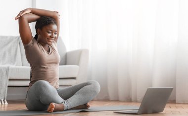 Ev sporu. Genç siyahi bayan dizüstü bilgisayarda ders seyrediyor oturma odasındaki yoga minderinde eğitim görüyor, kol kaslarını esnetiyor, gülümsüyor, spor yapıyor ve sağlıklı bir yaşam sürüyor.