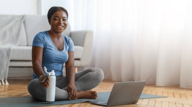 Spor kıyafetleri içinde spor kıyafetleri içinde ev ödevine hazır, Afrikalı kadın çevrimiçi derslerden ve ev içi fitness eğitiminden zevk alıyor, kameraya bakıyor ve gülümsüyor, uzay kopyaları yapıyor.