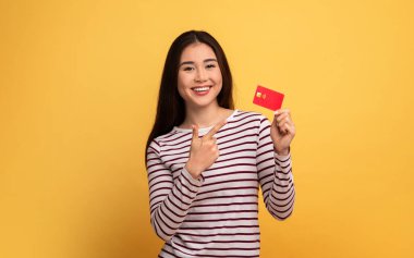 Asyalı, uzun saçlı genç bir kadın elinde kırmızı bir kart tutarken ışıl ışıl gülümsüyor. Parlak sarı bir arka plana karşı coşkuyla ona bakıyor. Heyecan ve pozitiflik taşıyor..