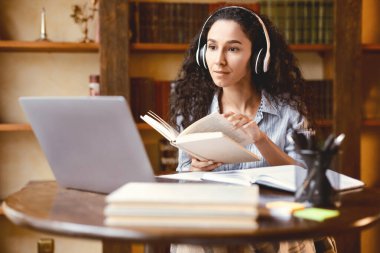 Sanal konferans. Kablosuz kulaklıklı genç bayanın portresi masada oturuyor, elinde kitap tutuyor ve okuyor, öğretmenle online ders alıyor ve dizüstü bilgisayara bakıyor. Evde çalışan bir kadın.