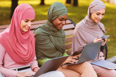 Modern Müslüman Öğrenci Kadınlar Açık Parkta Bench 'te oturan Laptop Bilgisayarları ve Akıllı Telefonla İnternet' te Öğrenim Amaçlı Öğreniyor. Uzak Eğitim Konsepti
