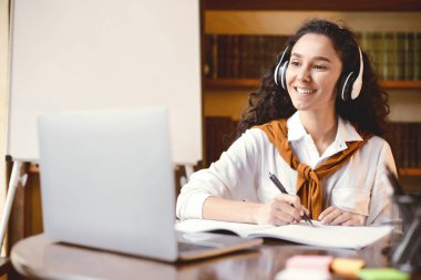 E-öğrenme kavramı. Pozitif neşeli genç kıvırcık kadın sanal konferans izliyor, dizüstü bilgisayara bakıyor, kalemle yazıyor ve defterine not alıyor, masada oturuyor. Kilitleme sırasında eğitim