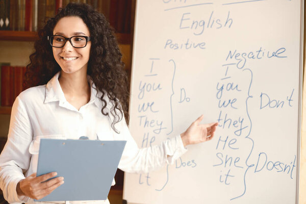 Training Concept. Portrait of cheerful positive female tutor wearing spectacles teaching English language, pointing at grammar rules on board with hand, looking and asking students, holding clipboard