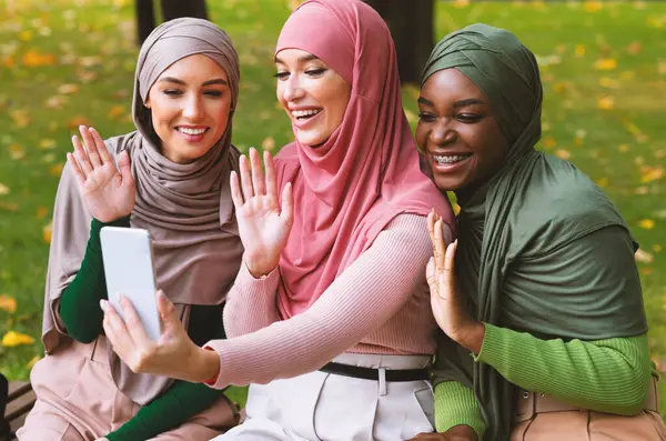 Üç farklı Müslüman kadın telefonda video görüşmesi yapıyor ve el sallayarak dışarıda parkta bankta oturarak iletişim kuruyor. Modern Uzaklık İletişim Kavramı