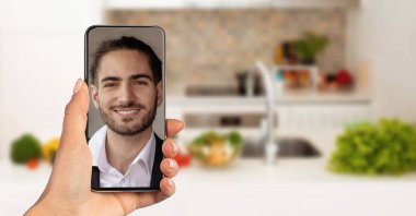 Bir kişi video görüşmesi sırasında gülümseyen bir adamı gösteren bir akıllı telefon tutuyor. Arka planda taze sebzeleri ve parlak bir atmosferi olan modern bir mutfak var..