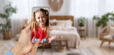 Bir kişi video görüşmesi sırasında gülümseyen bir çocuğu gösteren bir akıllı telefon tutuyor. Konforlu ev ortamında bitkiler ve yumuşak mobilyalarla dekore edilmiş bir yaşam alanı bulunuyor..
