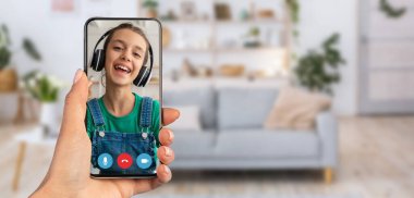 Genç bir çocuk akıllı telefondan yapılan bir video görüşmesiyle mutlu mesut meşgul oluyor. Dekor, arka planda bitki ve kanepe olan rahat bir oturma odası. Sıcak bir atmosfer yaratıyor..