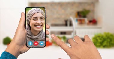Bir insan akıllı telefonuyla bir video görüşmesi yaparken gülümseyerek arkadaşça bir etkileşim sergiliyor. Sıcak mutfak arka planı sohbete sıcaklık katıyor..
