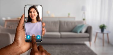 Samimi bir oturma odasında çevrimiçi video görüşmesi sırasında gülümseyen bir arkadaşını gösteren bir akıllı telefon tutan bir kişi. Sahne, evdeki modern iletişim ve bağlantıyı yakalıyor..