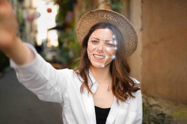 Genç bir kadın canlı bir sokak arasında selfie çekerken yüz tanıma teknolojisi kullanıyor. Modern teknoloji onun yüzünü bir şebeke örtüsüyle ön plana çıkarıyor, tanımlama gelişimini simgeliyor..