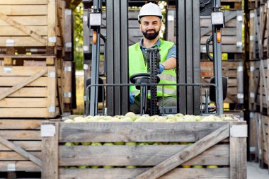 Meyve ve yiyecek dağıtımı. Kamyon elma dolu konteynırlarla dolu, pazara gönderilmeye hazır. Miğferli genç adam forklift kullanıyor, meyve kaldırıyor, arka planda bir sürü tahta kutu var.