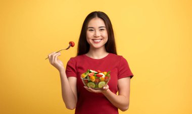 Genç bir Asyalı kadın elinde bir kase renkli salata tutarken gülümsüyor ve kiraz domatesi ile çatal kaldırıyor. Parlak sarı arka plan onun neşeli yüz ifadesini ve canlı yemeklerini vurguluyor..
