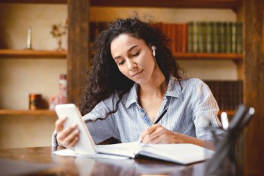 Günü planlıyorum. Kablosuz kulaklıklı genç bayan masada oturuyor, cep telefonunu tutuyor ve kullanıyor, kağıt defterine kalemle yazıyor, programı hakkında notlar alıyor, çevrimiçi kurslar izliyor.