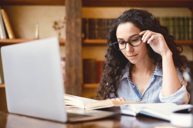 Eğitim ve Hobi Konsepti. Kağıt kitap okuyan, gözlüklere dokunan, evdeki ya da kütüphanedeki masada oturan, hikayeden zevk alan, masa başında dizüstü bilgisayar kullanan ve öğrenen genç bir kadının portresi.