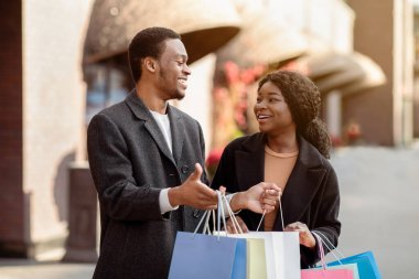 Aşk, tatil satışları, mağaza, perakende, tüketici. Binlerce yıllık neşeli Afro-Amerikan erkek ve bayanı bir sürü renkli alışveriş paketleriyle, adam alışveriş merkezinde alışveriş yaptıktan sonra çantalarında bir şeyler gösteriyor, bedava alan.