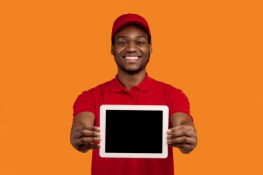 Teslimat Başvurusu. Afro-Amerikan kurye elinde boş ekran ile dijital tablet tutuyor ve onu kameraya gösteriyor, kırmızı başlık ve üniforma giyiyor, turuncu stüdyo arka planında izole edilmiş.