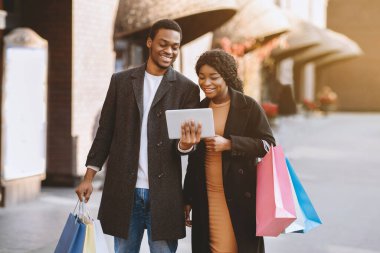Çevrimiçi bankacılık başvurusu, mağazalarda satış ve promosyon ilanı. Bir sürü renkli alışveriş torbası taşıyan mutlu Afro-Amerikan çifti tablete bakıyor, şehir merkezinde yürüyor, boş yer var.