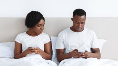 Genç siyahi kadın, erkek arkadaşını cep telefonuyla dinlerken akıllı telefon ekranına bakıyor. Afrika kökenli Amerikalı çift yatakta oturuyor ve cep telefonu kullanıyorlar. İlişkilerde güvensizlik
