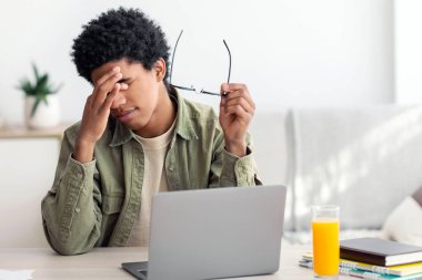 Black teenager feeling tired, rubbing irritated eyes, sitting at desk with laptop, exhausted from studying online. African American guy feeling fatigue from looking at screen, doing assignments on web