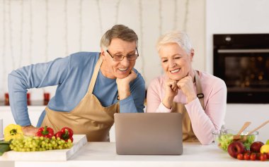 Retirement Hobby. Senior Couple Watching Online Cooking Course On Laptop Computer Sitting In Modern Kitchen At Home. Happy Retired Life, Elderly Spouses Watch Movie Preparing Dinner