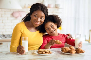Atıştırma zamanı. Mutlu siyah anne ve küçük kız ev yapımı pastadan zevk alıyor ve mutfakta süt içiyor, taze kruvasan ve kurabiyelerle bir ısırık alıyor, kameraya gülümsüyor, bedava uzay boşluğu.