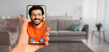 Samimi bir oturma odasındaki video görüşmesi için akıllı telefonu kullanan biri. Rahat mobilyalar ve sıcak ışıklandırma iletişim için hoş bir atmosfer yaratıyor..