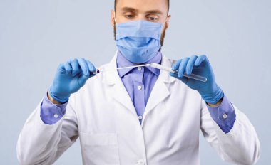 Yüz maskesi takmış tıp doktoru ve eldivenleri açık gri stüdyo arka planında PCR test tüpü tutuyor, panorama. Burun ve oral koronavirüs tanılama prosedürü için laboratuvar tüpü kullanarak pratisyen hekim deneyimi.