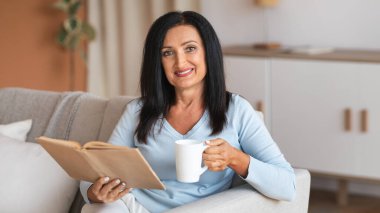 Evde vakit geçirmek. Yetişkin kadın portresi kitap okuyor ve elinde bir fincan kahve tutuyordu. Oturma odasındaki kanepede oturmuş hafta sonunun keyfini çıkarıyordu. Kameraya bakan kadın.