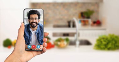 Çevrimiçi bir arama sırasında gülümseyen bir yüz gösteren bir akıllı telefon tutan bir kişi. Arka planda taze sebzelerle iyi aydınlatılmış bir mutfak var, evden bir anlık bir bağlantı yakalamış..