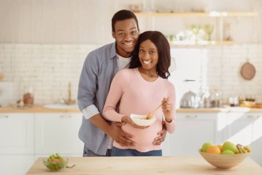 Sağlıklı hamilelik kavramı için diyet. Pozitif siyahi adam, sevgili müstakbel karısına mutfakta besleyici bir yemekle sarılıyor. Genç çift birlikte yemek pişiriyor ve evde sarılıyorlar.