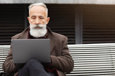 Yoğunlaşmış kır saçlı, uzun sakallı bir adam dışarıda oturuyor, dizüstü bilgisayar kullanıyor, kopyalama alanı kullanıyor. İş merkezinde bankta oturup klavyeyle yazı yazarken çevrimiçi çalışan yaşlı işadamı