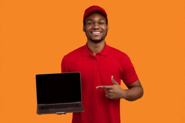 Siparişinizi izleyin. Sırıtkan siyah teslimatçının portresi elinde boş ekranla kırmızı başlık ve üniforma giyerek turuncu arka planda izole edilmiş boş ekranı işaret ediyor.