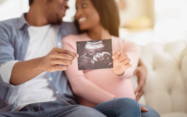 Pozitif siyah çift, doğmamış bebeklerinin ultrason fotoğraflarını evde gösteriyorlar. Ellerine odaklanmış durumdalar. Çocuğunun ultrasonuyla mutlu ve mutlu anılar yaratmayı bekleyen bir aile.