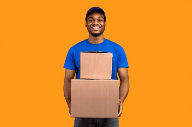 En iyi servis konsepti. Mavi şapkalı gülümseyen siyah teslimatçı ve elinde turuncu stüdyo arka planında izole edilmiş karton kutular tutan biçimsiz adam. Afro-Amerikan kurye paketleri taşıyor.